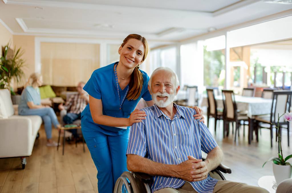 A smiling healthcare worker in blue scrubs stands beside an elderly man in a wheelchair. They are part of the NHTD Waiver Program, fostering independence in a bright, comfortable room where other individuals engage in warm conversations.